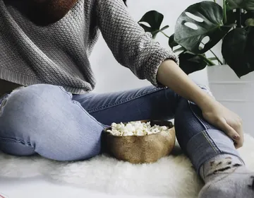 Personne assise sur un tapis en pull gris et jean, un bol de pop-corn en bois posé à côté d'elle